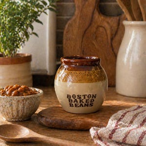 May include: A ceramic jar with the text "BOSTON BAKED BEANS" in black lettering. The jar is two-toned, with a brown top and a beige base. A small bowl of baked beans, a wooden spoon, and a striped cloth are also visible.