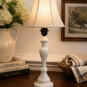 May include: A table lamp with a white, fluted, pedestal-style base featuring ornate, carved-look detailing at the top and bottom. The lamp is topped with a flared, off-white fabric shade and a small, round finial, emitting a soft, warm glow.