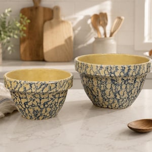 May include: Two ceramic mixing bowls featuring a cream-colored interior and a blue and white sponge-painted exterior pattern. The bowls have a wide, thick rim and a rounded base, showcasing a speckled, textured appearance on the outer surface.