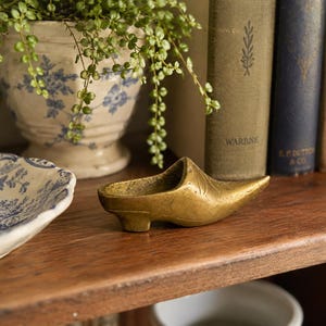 May include: A small, hollow brass figurine shaped like a pointed-toe shoe or clog. The object has a metallic gold finish with subtle etched line details on the upper surface and a low, block-style heel. It is a singular, decorative metal item.