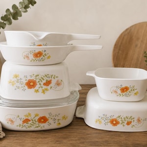 May include: A collection of white ceramic cookware with floral designs. The set includes various sizes of casserole dishes, a saucepan, and a skillet. The floral pattern features orange, yellow, and blue flowers with green leaves. The cookware is stacked on a wooden surface.