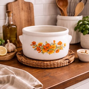 May include: White ceramic casserole dish with orange and yellow floral design, set on a round woven placemat. The dish has two handles and is displayed on a wooden countertop in a kitchen setting.