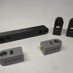 Op de afbeelding: Een assortiment zwart en grijs 3D-geprinte onderdelen. De onderdelen omvatten een langwerpig rechthoekig stuk, verschillende klemachtige stukken en blokken met het woord "RAM" in rood. Sommige stukken hebben goudkleurige inzetstukken.