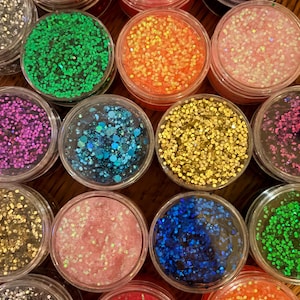 Peut inclure: Une collection de petits pots en plastique transparent remplis de paillettes de différentes couleurs. Les couleurs des paillettes comprennent le rouge, le rose, le bleu, le vert, l'orange, le violet et l'or. Les pots sont disposés en grille, mettant en valeur la variété de paillettes.