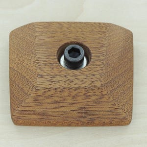 May include: A square, wooden furniture component with a dark brown finish. A black bolt is centered in the wood. The edges of the wood are rounded. The wood grain is visible.