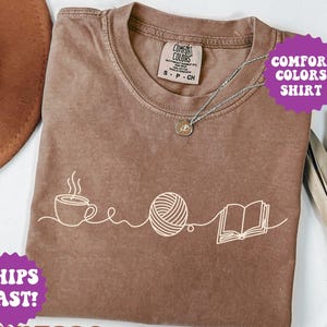 May include: Espresso-colored Comfort Colors shirt with a white graphic of a coffee cup, yarn ball, and open book. The shirt has a crew neck and short sleeves. A silver necklace with a pendant is visible.