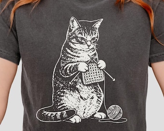 May include: Dark gray t-shirt featuring a white illustration of a cat knitting. The cat wears glasses and holds knitting needles, with a ball of yarn nearby. The word "PEPPER" is printed at the bottom of the shirt.