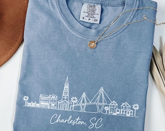 May include: A blue jean Comfort Colors t-shirt with a white line drawing of the Charleston, SC skyline. The shirt has a crew neck and short sleeves. A gold necklace with a pendant is draped across the shirt.