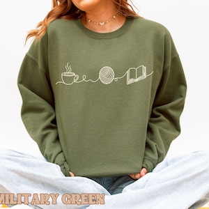 May include: Military green sweatshirt with a white line drawing of a coffee cup, yarn ball, and open book. The text "MILITARY GREEN" is at the bottom of the image. The sweatshirt is a solid color and has long sleeves.
