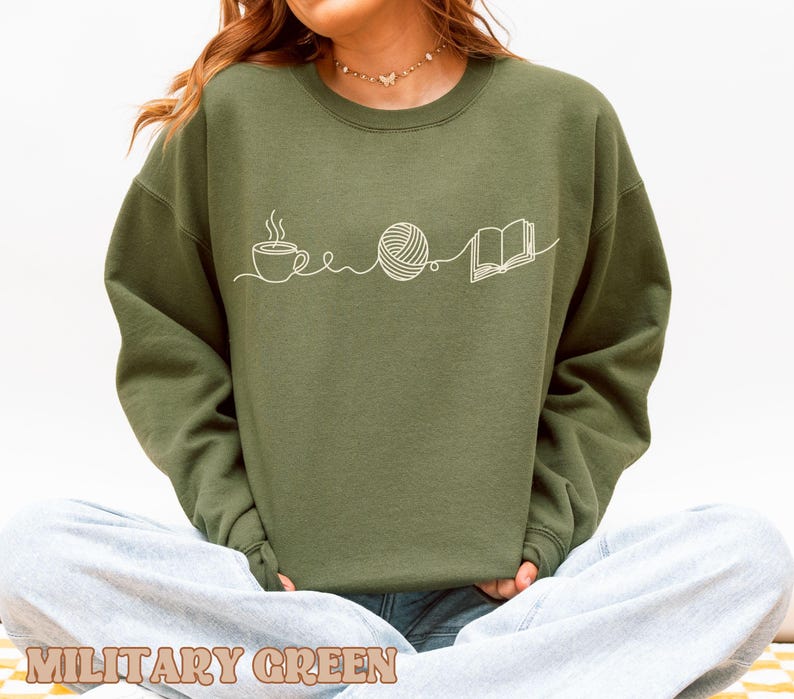 May include: Military green sweatshirt with a white line drawing of a coffee cup, yarn ball, and open book. The text "MILITARY GREEN" is at the bottom of the image. The sweatshirt is a solid color and has long sleeves.