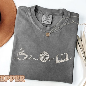 May include: A gray t-shirt with a white graphic of a coffee cup, yarn ball, and book. The word "PEPPER" is at the bottom. A silver necklace with a round pendant is on the shirt.
