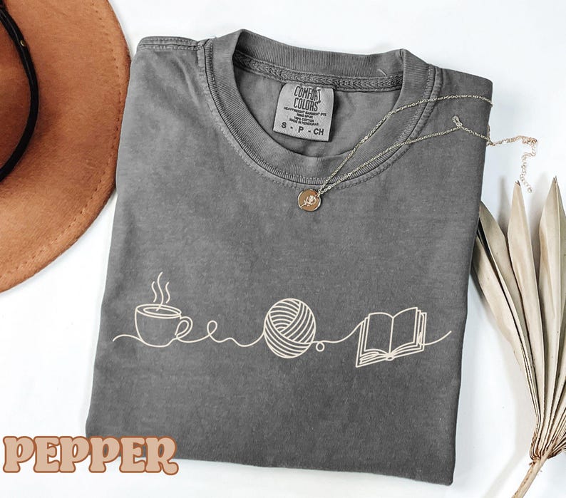 May include: A gray t-shirt with a white graphic of a coffee cup, yarn ball, and book. The word "PEPPER" is at the bottom. A silver necklace with a round pendant is on the shirt.