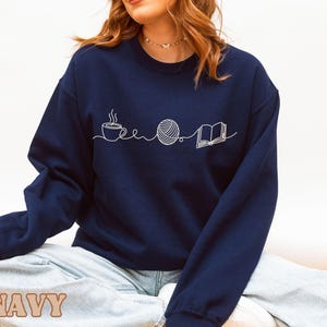 May include: Navy blue sweatshirt with a white line drawing of a coffee cup, a ball of yarn, and an open book. The word "NAVY" is printed in a tan, blocky font at the bottom of the image. The sweatshirt is a crew neck style.