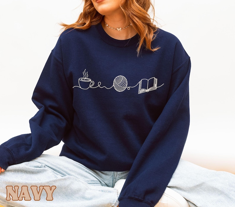 May include: Navy blue sweatshirt with a white line drawing of a coffee cup, a ball of yarn, and an open book. The word "NAVY" is printed in a tan, blocky font at the bottom of the image. The sweatshirt is a crew neck style.