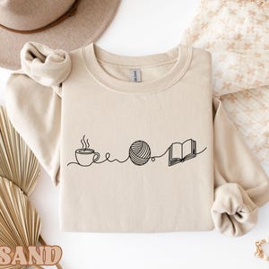 May include: A beige sweatshirt features a black line drawing of a coffee cup, a ball of yarn, and an open book. The design suggests a cozy theme, perfect for crafters and book lovers. The sweatshirt has long sleeves and a crew neck.