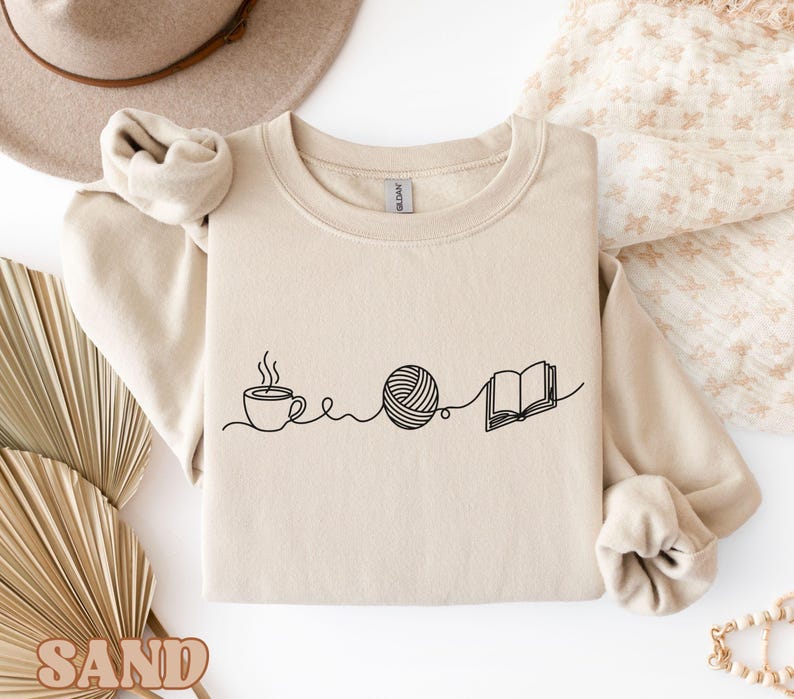 May include: A beige sweatshirt features a black line drawing of a coffee cup, a ball of yarn, and an open book. The design suggests a cozy theme, perfect for crafters and book lovers. The sweatshirt has long sleeves and a crew neck.