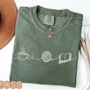 May include: Moss green t-shirt with a white graphic of a coffee cup, yarn ball, and open book. The shirt has a crew neck and short sleeves. A gold necklace is draped across the shirt. The word "MOSS" is printed in brown at the bottom.