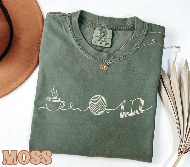 May include: Moss green t-shirt with a white graphic of a coffee cup, yarn ball, and open book. The shirt has a crew neck and short sleeves. A gold necklace is draped across the shirt. The word "MOSS" is printed in brown at the bottom.