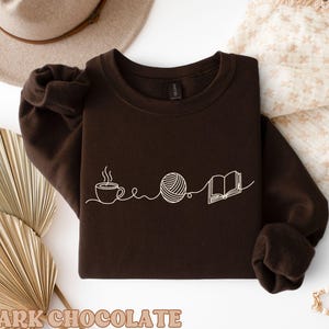May include: Dark chocolate brown sweatshirt with a white line drawing of a coffee cup, yarn ball, and open book. The words "DARK CHOCOLATE" are printed at the bottom of the image. The sweatshirt has long sleeves and a crew neck.