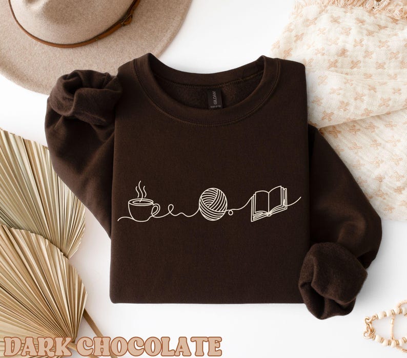 May include: Dark chocolate brown sweatshirt with a white line drawing of a coffee cup, yarn ball, and open book. The words "DARK CHOCOLATE" are printed at the bottom of the image. The sweatshirt has long sleeves and a crew neck.