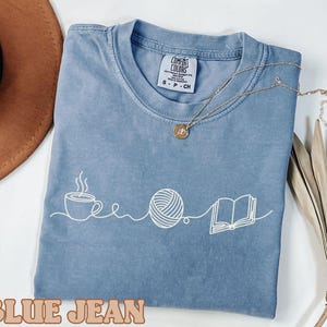 May include: A blue jean colored t-shirt with a white graphic of a coffee cup, a ball of yarn, and an open book. The shirt has a crew neck and short sleeves. A gold necklace is draped on the shirt.