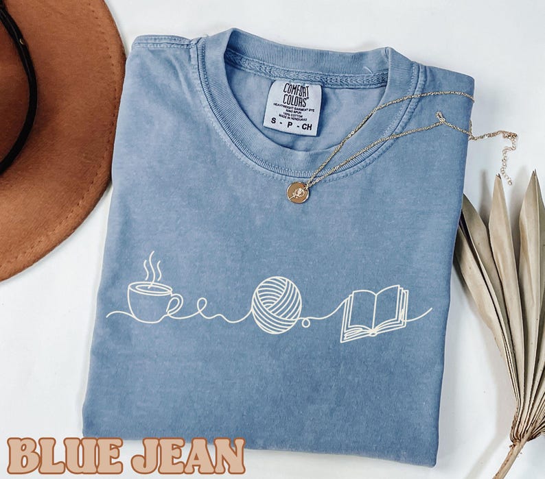 May include: A blue jean colored t-shirt with a white graphic of a coffee cup, a ball of yarn, and an open book. The shirt has a crew neck and short sleeves. A gold necklace is draped on the shirt.
