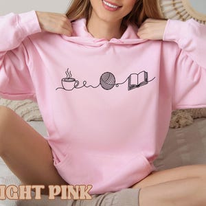May include: Light pink hoodie with a black line drawing of a coffee cup, yarn ball, and open book. The text "LIGHT PINK" is at the bottom of the image. The hoodie has a front pocket and a hood.