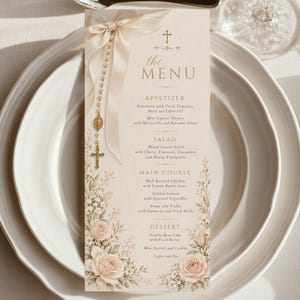 May include: A vertical menu card featuring a cream background, a cross icon, and the text "the MENU". The design includes a printed illustration of a rosary with a beige ribbon, pink roses, and greenery. It is placed on a white dinner plate.