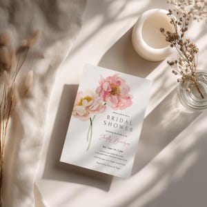 Pu&ograve; includere: Un invito per un addio al nubilato con peonie rosa e gialle ad acquerello. L'invito include il testo "Bridal Shower" e i dettagli dell'evento. Sono presenti anche un piccolo vaso di vetro con fiori secchi, una candela bianca e fiori secchi decorativi.