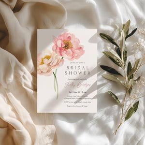 Pu&ograve; includere: Un invito per un addio al nubilato con peonie rosa ad acquerello. L'invito include il testo "BRIDAL SHOWER" e i dettagli dell'evento. &Egrave; posto su una superficie bianca, accompagnato da rami di ulivo e tessuto morbido.