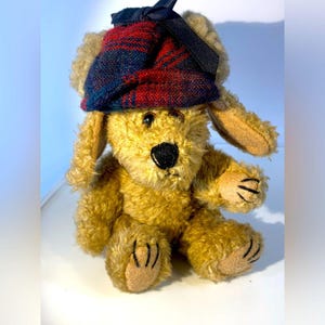 Pode incluir: Um brinquedo de pelúcia de cachorro cor de bronzeado com orelhas caídas e um nariz preto. O cachorro usa um chapéu xadrez em vermelho, azul marinho e preto, com um laço preto. O brinquedo está sentado, com as patas visíveis.
