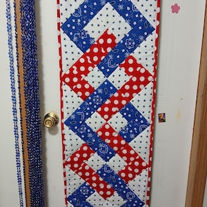 Puede incluir: Un tapiz de pared acolchado de temática patriótica. La pieza rectangular presenta un patrón de lunares rojos, paisley azul y estrellas blancas. El diseño está enmarcado con un borde rojo y colgado con una barra de madera y pinzas para la ropa.