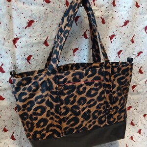 May include: A tote bag with a leopard print pattern in brown and black. The bottom of the bag is a solid dark brown. The bag has two handles and a small pocket with a red trim.