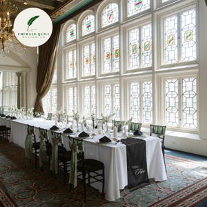 May include: A long banquet table set for a formal event with white tablecloths, black napkins, and sage green chair sashes. A black table runner features the text Table Five. The room has tall, multi-paned windows with stained glass accents.