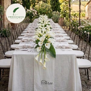 May include: A long, white linen-covered table set for a formal outdoor event. The table is decorated with a floral centerpiece of white calla lilies and greenery. The word "Three" is printed on the table runner. Chairs with white cushions are arranged around the table.