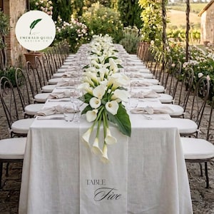 May include: A long rectangular dining table covered with a white linen tablecloth, featuring a central runner with the text TABLE Five. A floral arrangement of white calla lilies runs down the center, flanked by place settings with folded napkins.