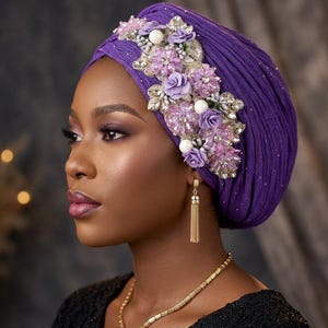 May include: A deep purple turban featuring a textured fabric base adorned with a decorative side panel of lavender fabric flowers, white faux pearls, and clear crystal embellishments. The headpiece is designed with intricate pleating throughout.