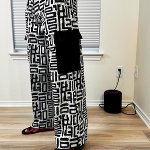 Op de afbeelding: Zwarte en witte broek met patroon en een zwarte zak. De broek heeft een geometrisch ontwerp en wordt gedragen met een witte top en sandalen. Het patroon bestaat uit zwarte vormen op een witte achtergrond.