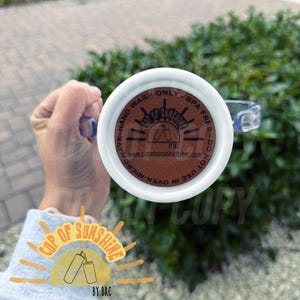 May include: White mug with a clear handle, featuring the text "Cup of Sunshine" and "Hand Wash Only". The bottom of the mug has a brown circular design with text and a logo. The mug is held against a green background.
