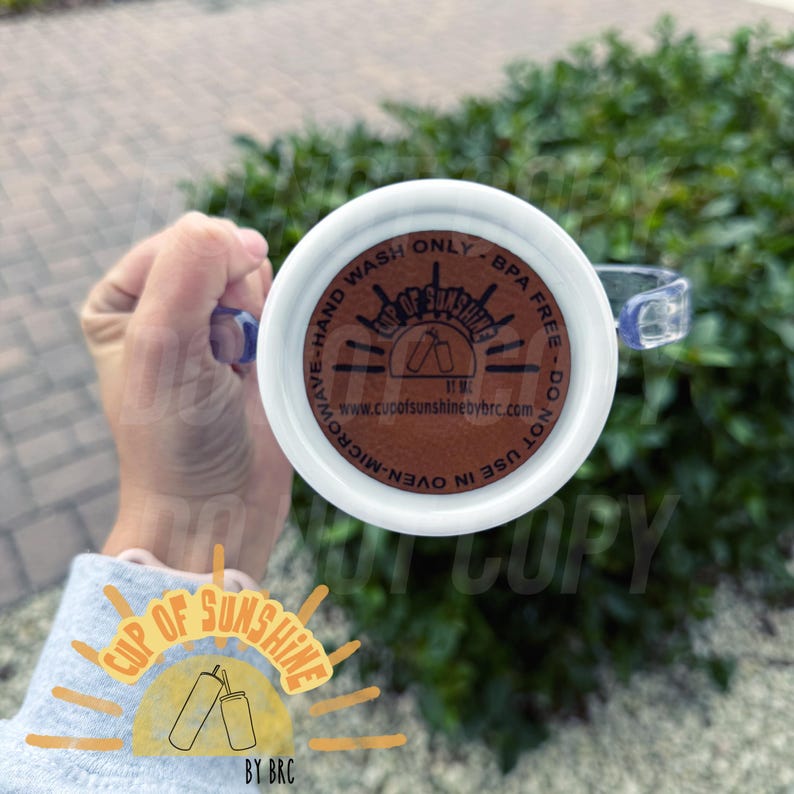 May include: White mug with a clear handle, featuring the text "Cup of Sunshine" and "Hand Wash Only". The bottom of the mug has a brown circular design with text and a logo. The mug is held against a green background.