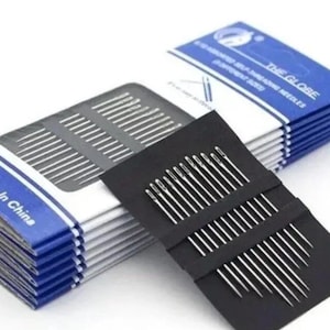 May include: A stack of needle packets with blue and white labels, and the text "THE GLOBE". The packets are labeled "Made in China". Several silver sewing needles are visible in a black holder.