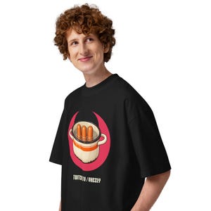 Può includere: Una t-shirt nera a maniche corte con la grafica di una pentola bianca con una striscia arancione contenente tre wurstel in acqua, su una forma a mezzaluna rosa. Il testo TWITCH.TV/VHEZLY è stampato in bianco sotto la grafica.