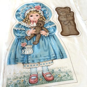May include: A fabric panel featuring a vintage illustration of a girl in a blue dress holding a teddy bear. The girl wears a bonnet and carries a floral purse. Additional teddy bear and bonnet illustrations are also present.