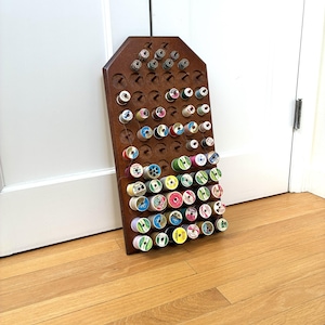 May include: A dark brown wooden wall-mounted thread rack with a pointed top, featuring numerous pegs holding various spools of sewing thread in assorted colors. The rack is organized with multiple rows of pegs, some occupied by colorful thread spools.