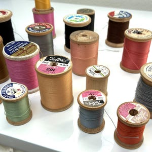 Könnte beinhalten: Eine Sammlung von Nähgarnspulen aus Holz in verschiedenen Farben wie Rosa, Blau, Braun, Grau und Orange. Jede Spule hat oben ein rundes Papieretikett mit Markennamen wie Talon und Coats & Clark, auf dem Text und Preisangaben zu sehen sind.