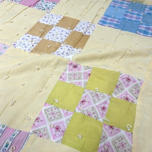 May include: A handmade patchwork quilt with a light yellow background. The quilt features squares of various fabrics in shades of pink, green, blue, and tan. Some squares have floral patterns, while others are solid colors. The quilt is a mix of textures and colors.