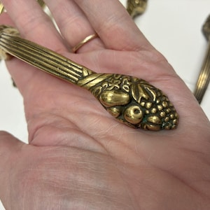 Op de afbeelding: Decoratieve messing lade-trekkers met een fruit- en bladeren ontwerp. De trekkers hebben een geribbelde handgreep en een decoratief uiteinde met druiven, bessen en bladeren. Het messing heeft een warme, verouderde patina.