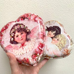 May include: Two heart-shaped decorative tins. One tin features a cherub with a floral crown and wings, surrounded by pink flowers. The second tin has two cherubs. Both tins have a pink and white colour scheme.