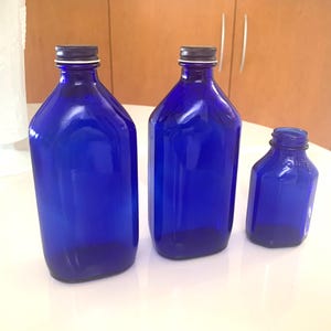 May include: Three cobalt blue glass bottles of varying sizes. The bottles have a rectangular shape with rounded shoulders and a dark metal cap. The bottles are empty and sitting on a white surface.