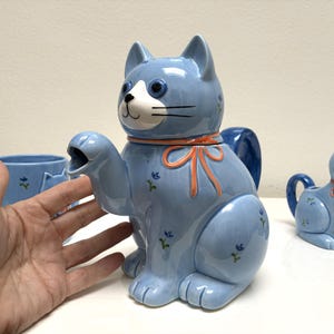 Könnte beinhalten: Hellblaue Keramik-Teekanne in Katzenform mit orangefarbener Schleife und floralen Akzenten. Die Katze hat ein weißes Gesicht, schwarze Schnurrhaare und blaue Augen. Eine passende Tasse und ein Sahnekännchen sind ebenfalls zu sehen.